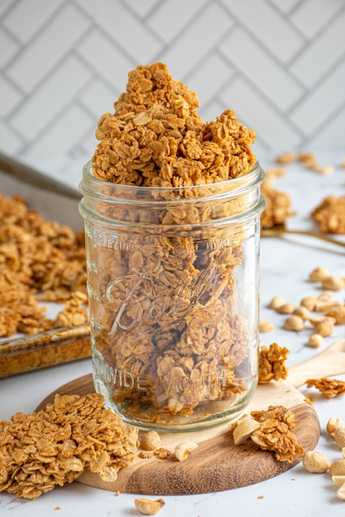 A mason jar full of large chunks of peanut butter granola surrounded by granola clusters and peanuts.