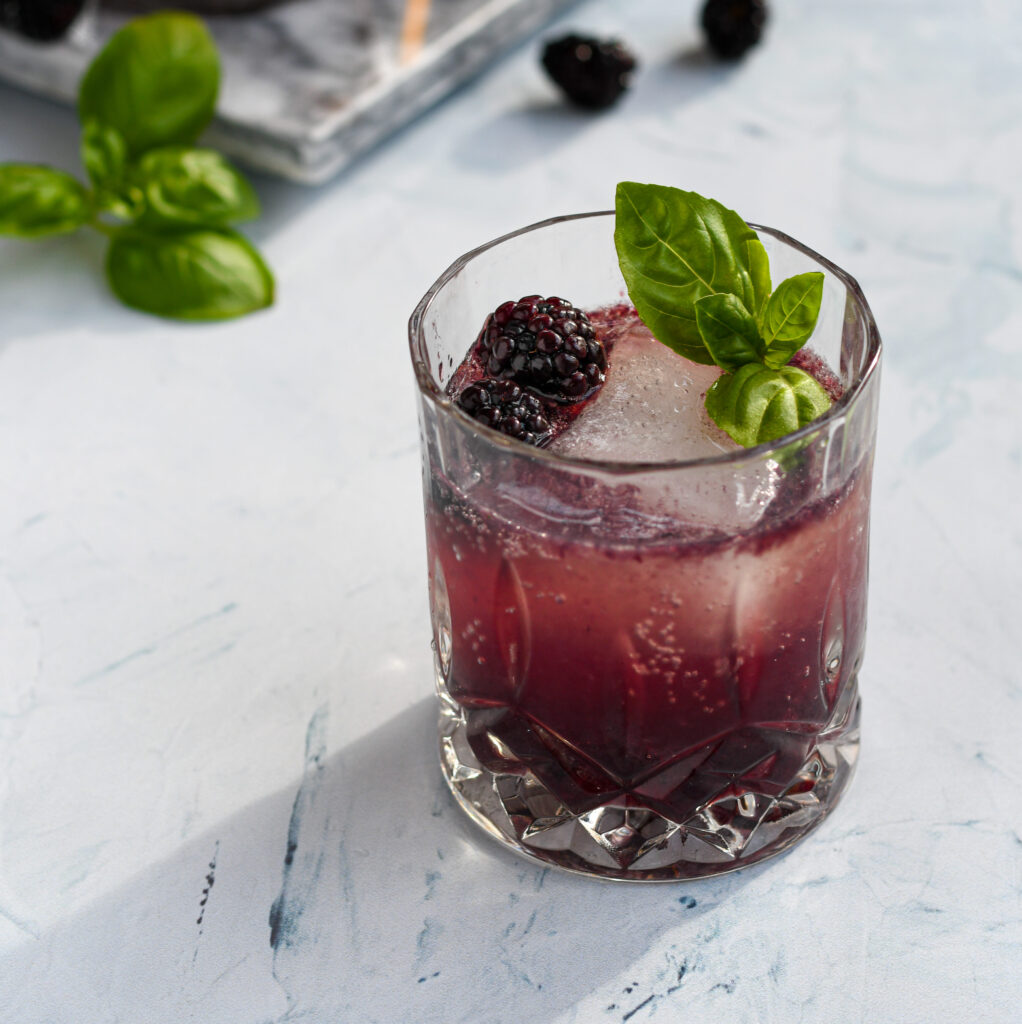 an old fashioned glass filled with a purple blackberry bourbon smash garnished with a large ice cube, blackberries, and basil