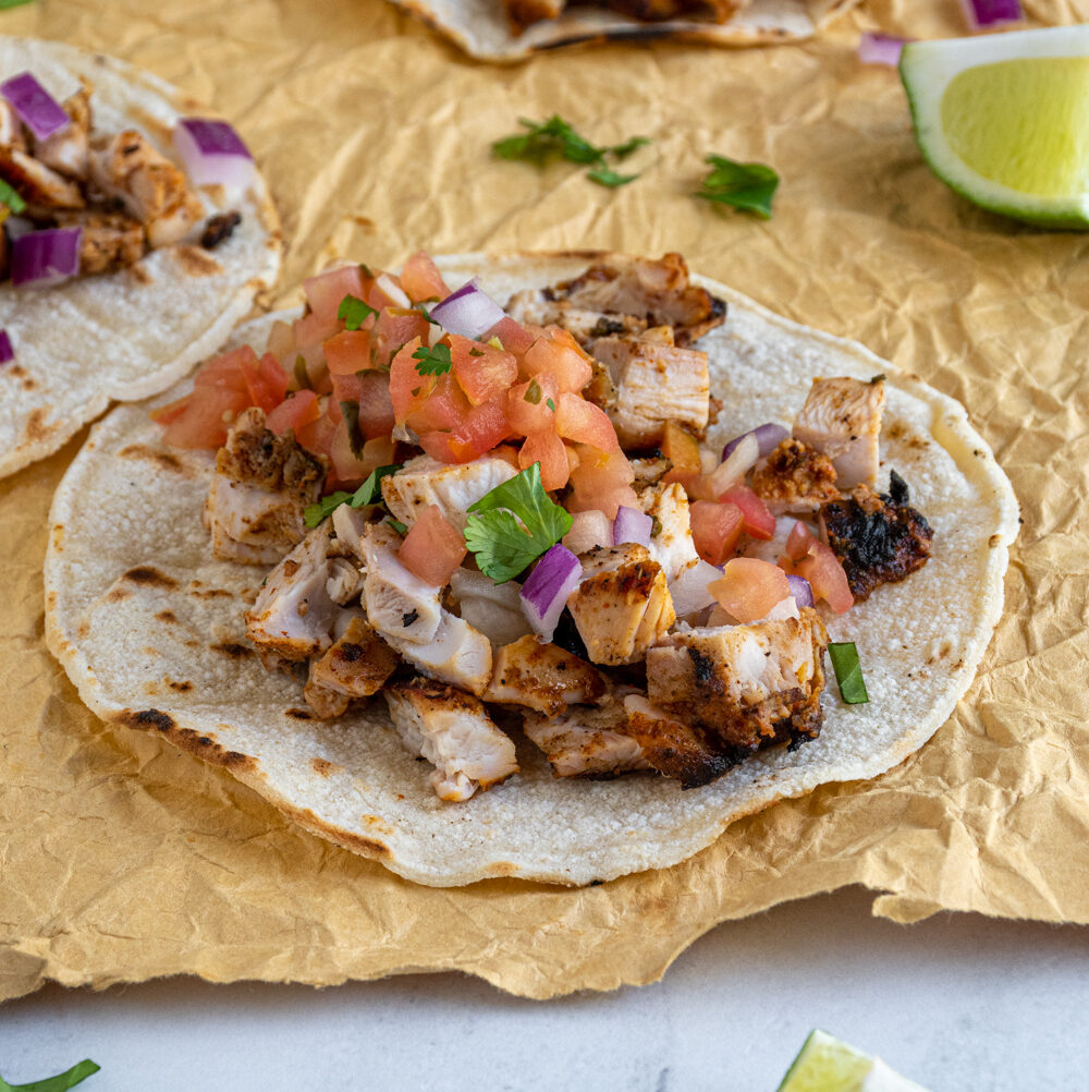 A grilled chicken street taco laid out on a table top filled with chicken and topped with salsa, onions, and cilantro.