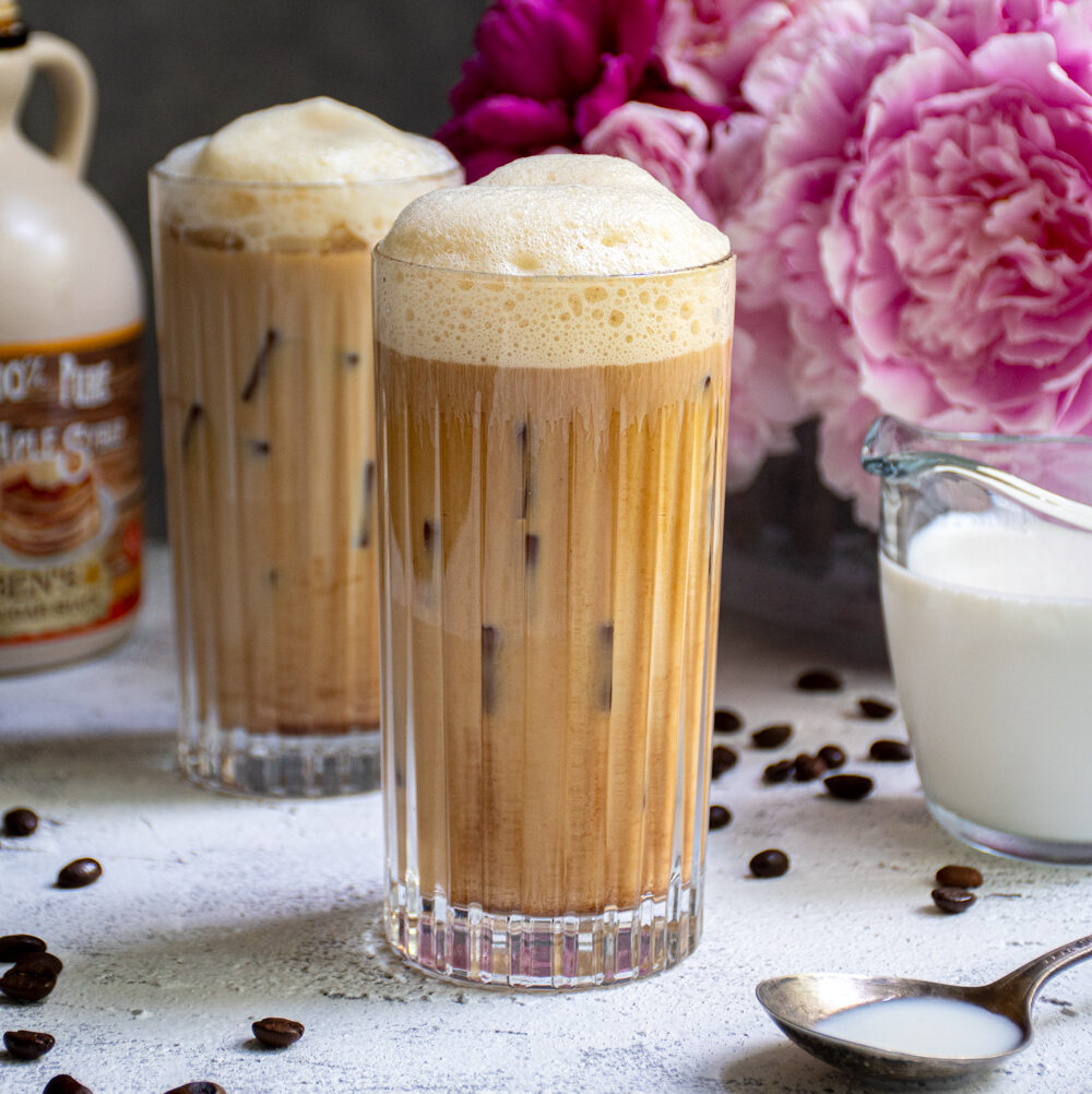 A glass filled with ice and salted maple cold brew coffee sitting on a table top next to another drink and a vase of pink flowers.