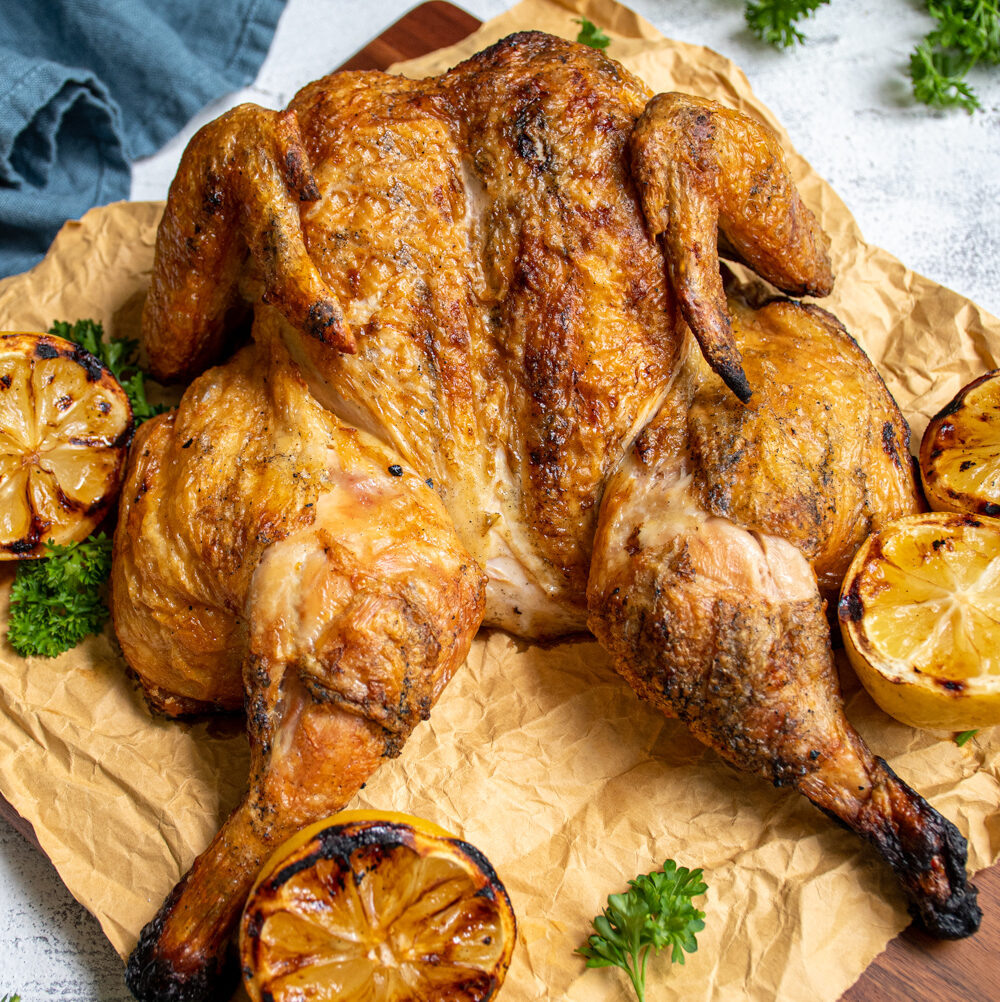 Grilled golden brown spatchcocked chicken sitting on butcher paper and a cutting board next to grilled lemons and parsley.