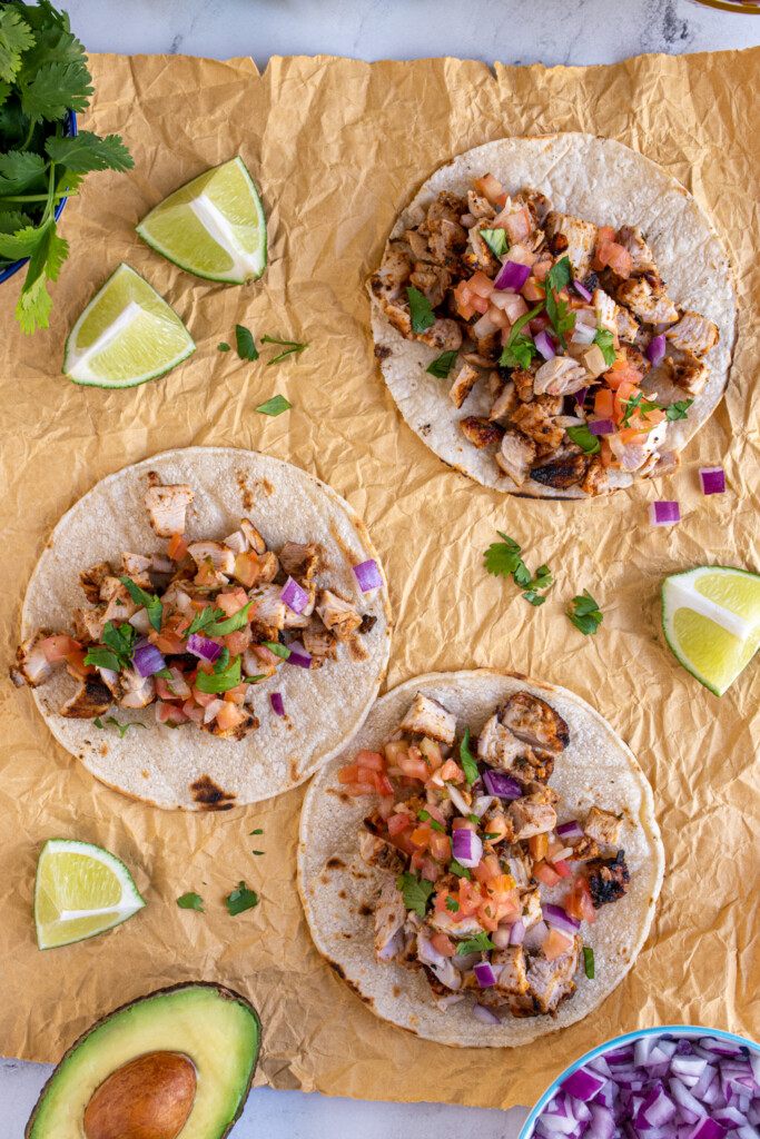 Overhead view of 3 corn tortillas filled with grilled chicken street taco, with garnishes like limes and cilantro.