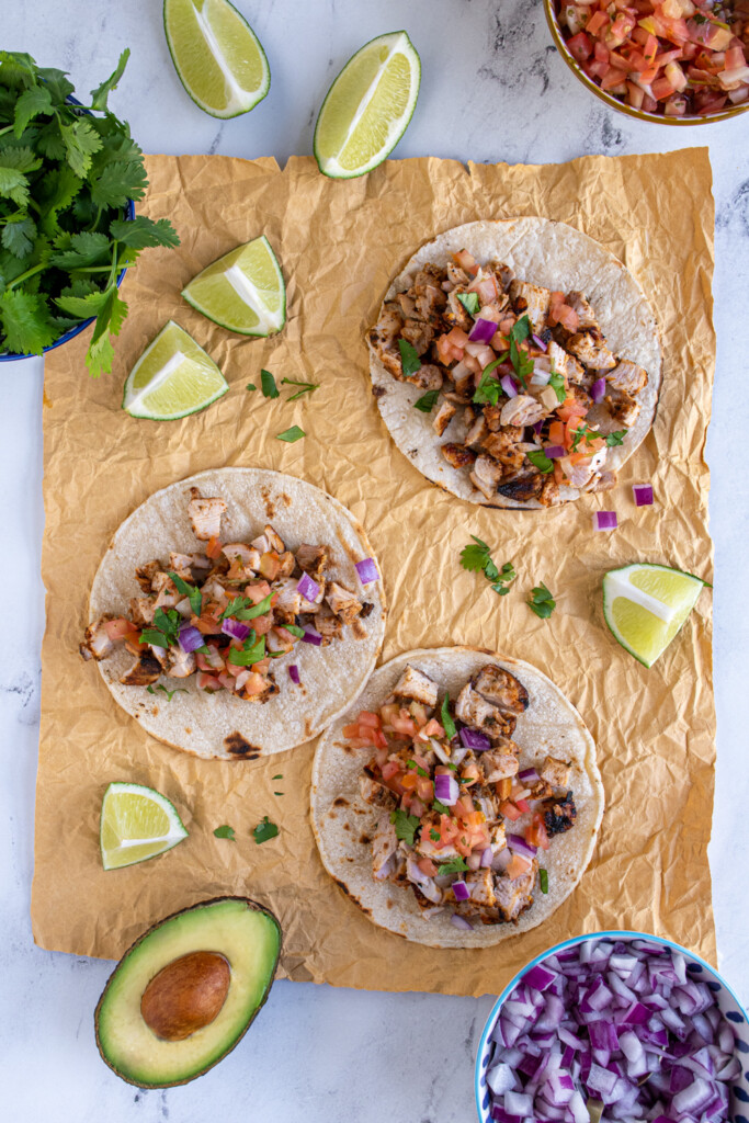 Overhead view of 3 grilled chicken street tacos garnished with salsa, onion, and cilantro and sitting next to lime wedges.