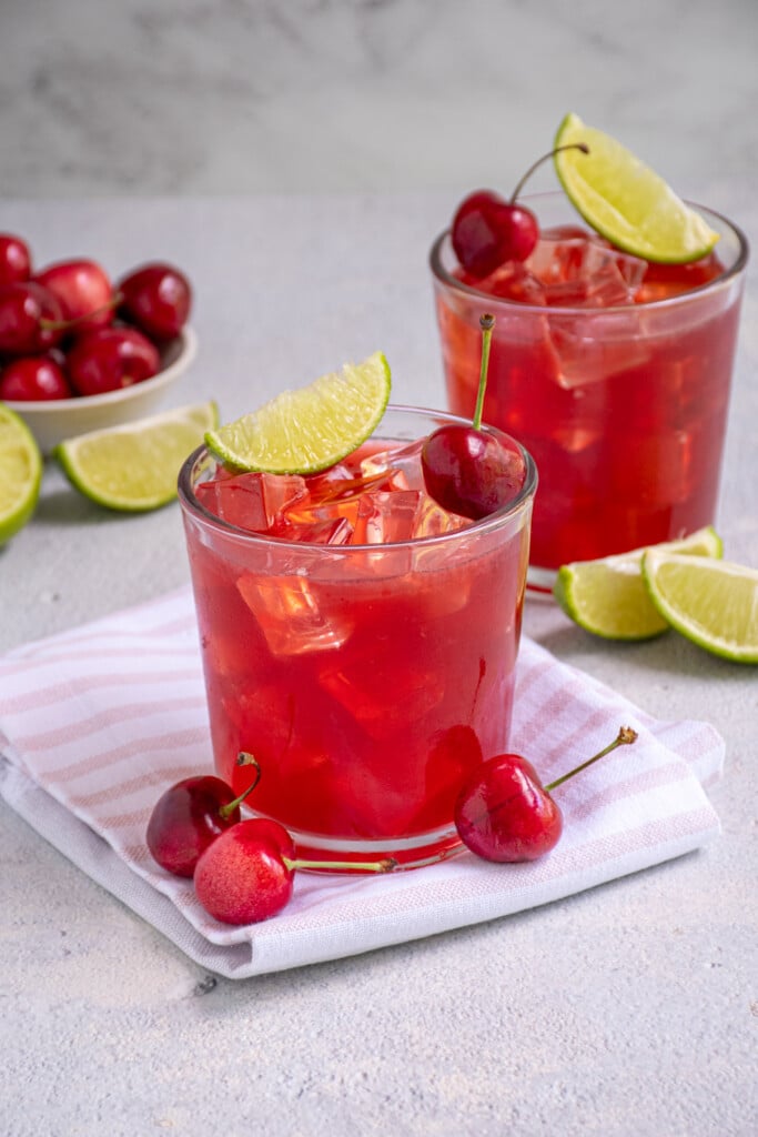 A glass filled with ice and a fresh cherry margarita garnished with a fresh cherry and a lime wedge.