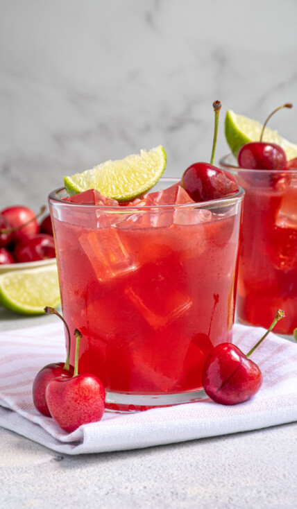 A glass filled with ice and bright red cherry margarita topped with a fresh cherry and lime wedge.