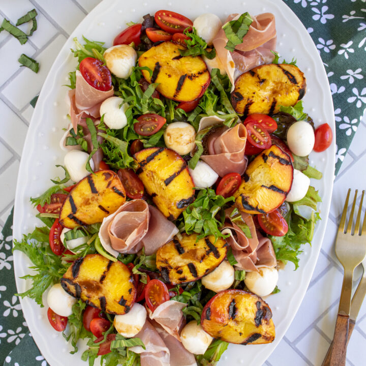 Overhead view of a large white platter with Grilled Peach Caprese Salad garnished with fresh basil leaves.