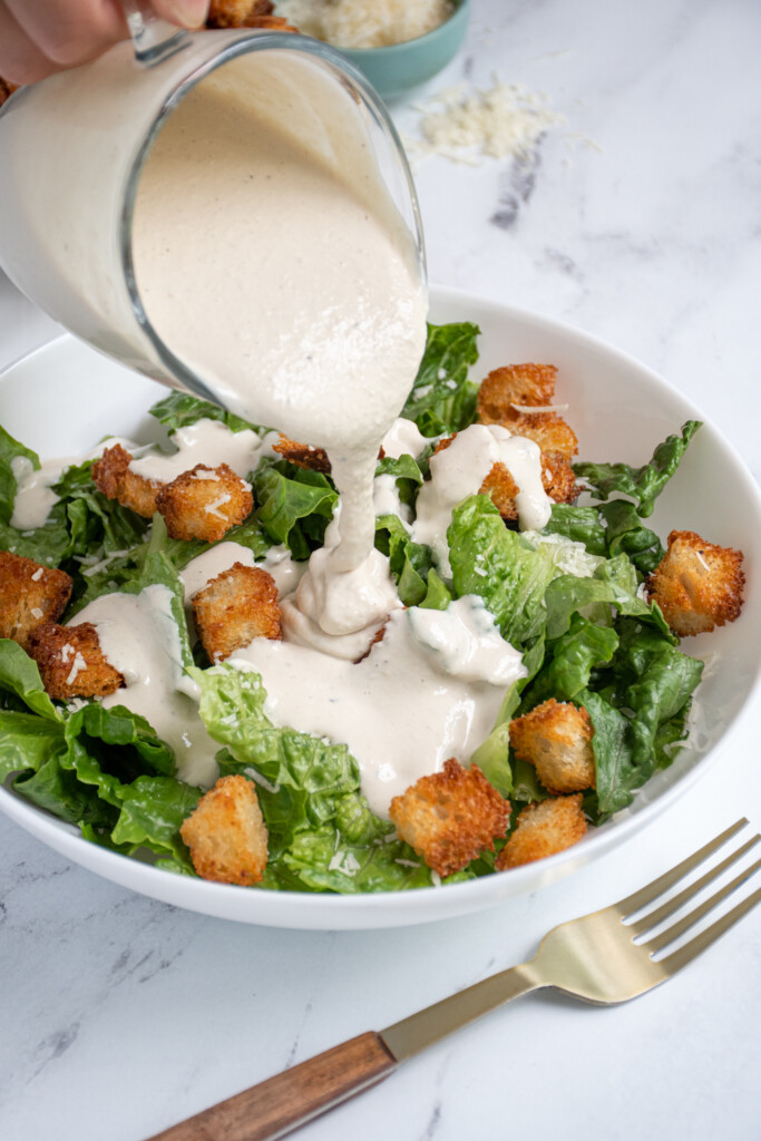 Pouring Greek Yogurt Caesar Dressing overtop of bed of lettuce and croutons.