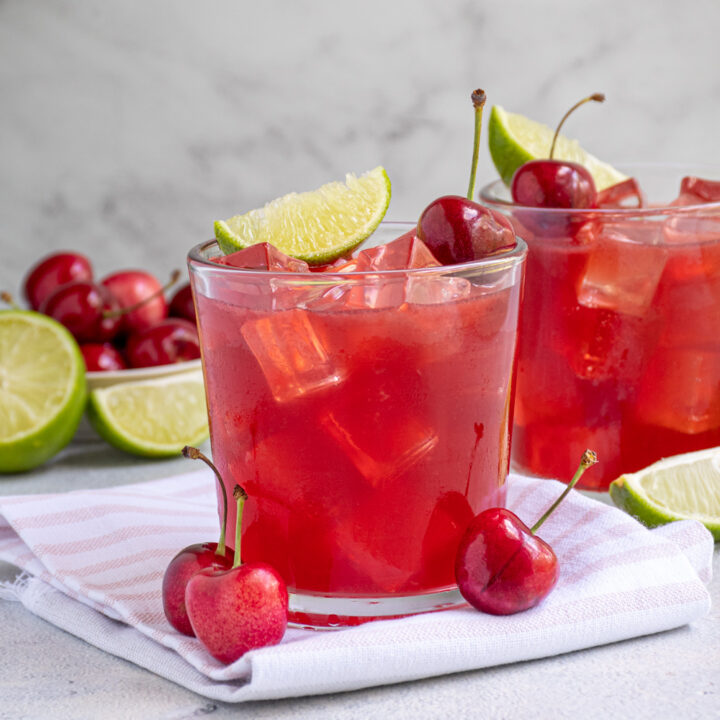 Glass filled with ice and a bright red cherry margarita topped with a fresh cherry and lime wedge.