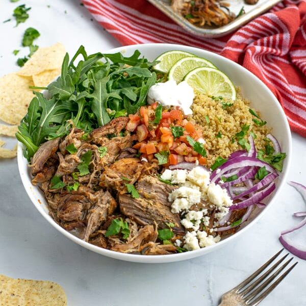 a large white bowl full of leftover carnitas, quinoa, arugula, and fresh toppings like salsa and sour cream