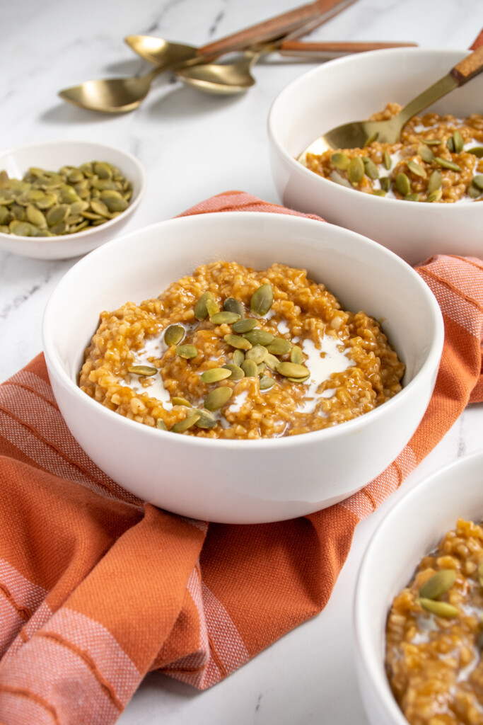White bowl filled with orange hued pumpkin spice steel cut oats topped with cream and pumpkin seeds.