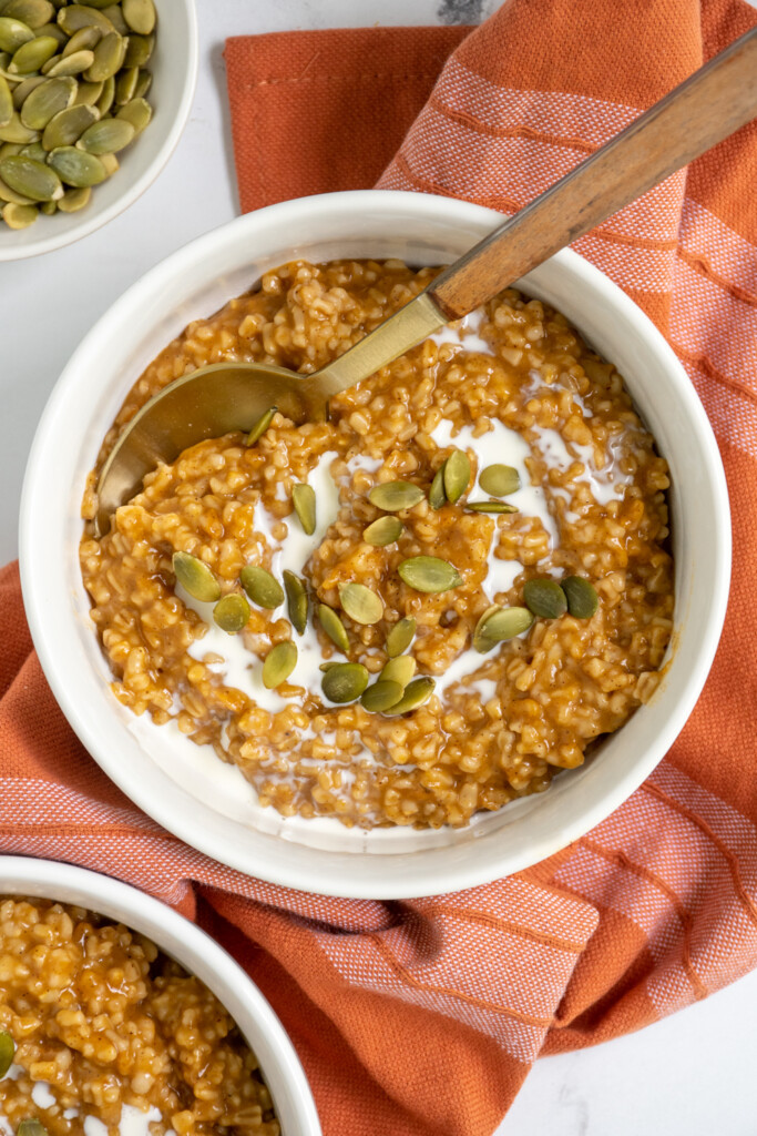Overhead view of a white bowl with a gold spoon on top of orange colored pumpkin spice steel cut oats garnished with cream and pumpkin seeds.