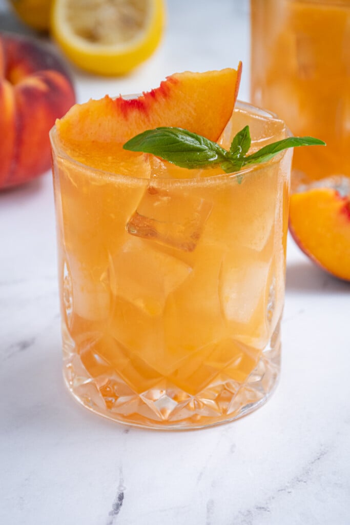 Close up of a whiskey glass with an orange colored Bourbon Peach Smash with a peach slice and basil as a garnish.