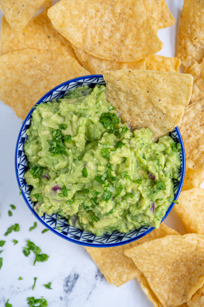 A blue bowl filled with simple guacamole surrounded by tortilla chips and garnished with cilantro.
