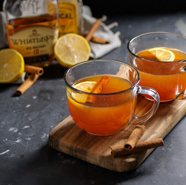 Two clear mugs on a wooden cutting board against a black background filled with hot toddy cocktail and garnished with cinnamon sticks and a lemon wedge.