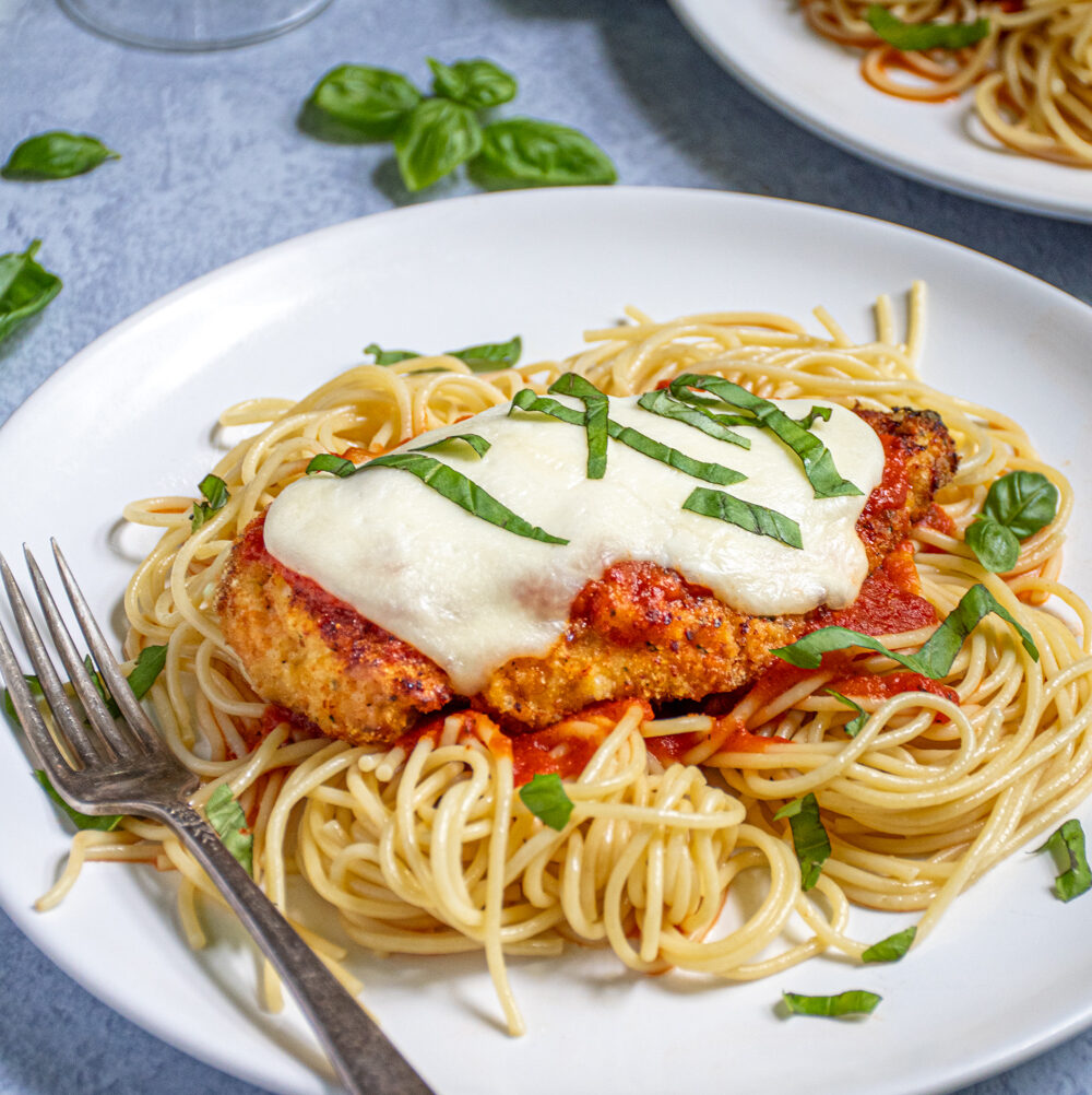 A white plate piled high with spaghetti topped with an Oven Baked Chicken Parmesan topped with melted mozzarella and garnished with pieces of fresh basil.