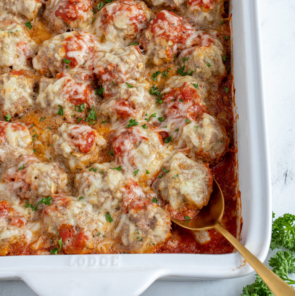 White casserole dish with baked turkey meatballs in sauce covered with cheese and fresh parsley.