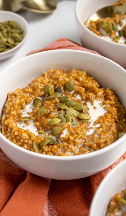 Close up of a white bowl filled with orange hued pumpkin spice steel cut oats that were made in the pressure cooker and topped with cream and pumpkin seeds.