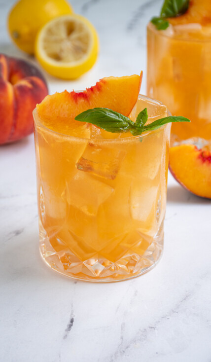 A glass full of Bourbon Peach Smash an orange colored cocktail garnished with a peach slice and basil leaves.