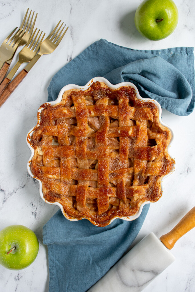 Overhead view of a homemade apple pie on top of a blue towel next to some forks and apples.