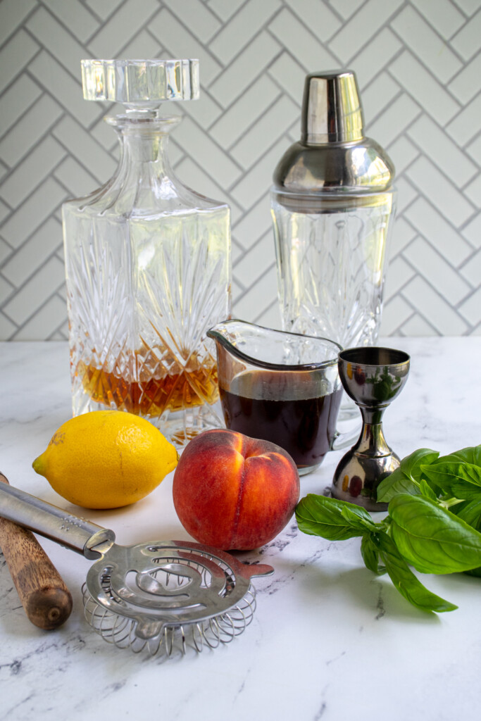 Ingredients to make a bourbon peach smash including equipment like a bar shaker and strainer.