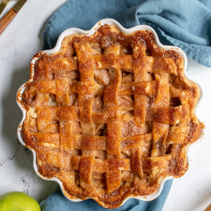Overhead view of a freshly baked apple pie laying on a blue towel next to some forks and some apples.