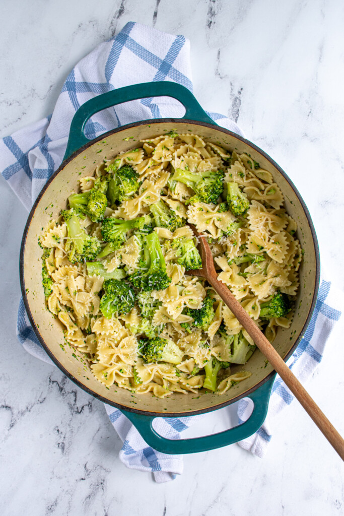 Overhead view of a dutch oven pit filled with bowties and broccoli pasta dish with a wooden serving spoon.