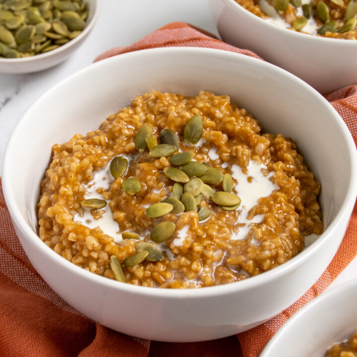 Close up of a white bowl filled with instant pot pumpkin oatmeal garnished with cream and pumpkin seeds.