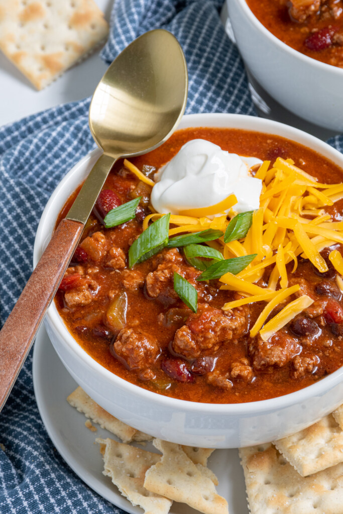 Close up of a bowl of chili in a white bowl the chili has chances of ground beef, peppers, and beans and is topped with sour cream, cheese, and green onion.
