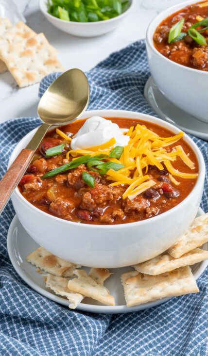 Bowl of classic chili with ground beef and beans topped with sour cream, cheese, and green onion with saltine crackers.