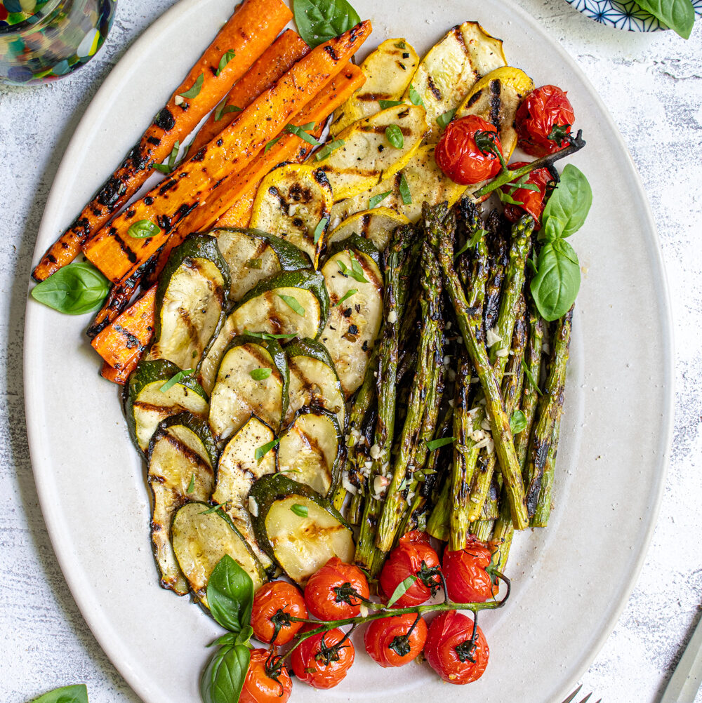 Overhead view of a white platter filled with marinated grilled veggies including zucchini, asparagus, carrots, and tomatoes.
