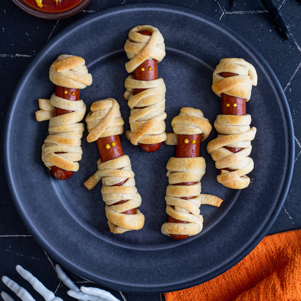 On a black background on a black plate sits five mummy hot dogs wrapped in golden brown dough and mustard dot eyes.