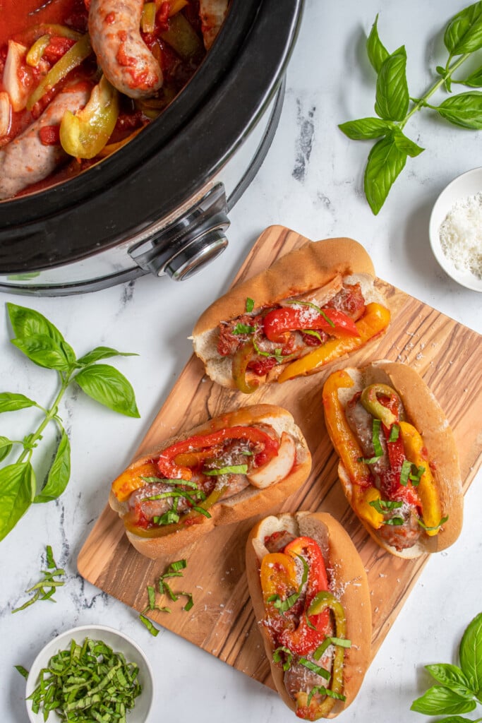 Overhead view of a bunch of buns filled with Italian sausages and peppers sitting next to a crockpot with more sausage.