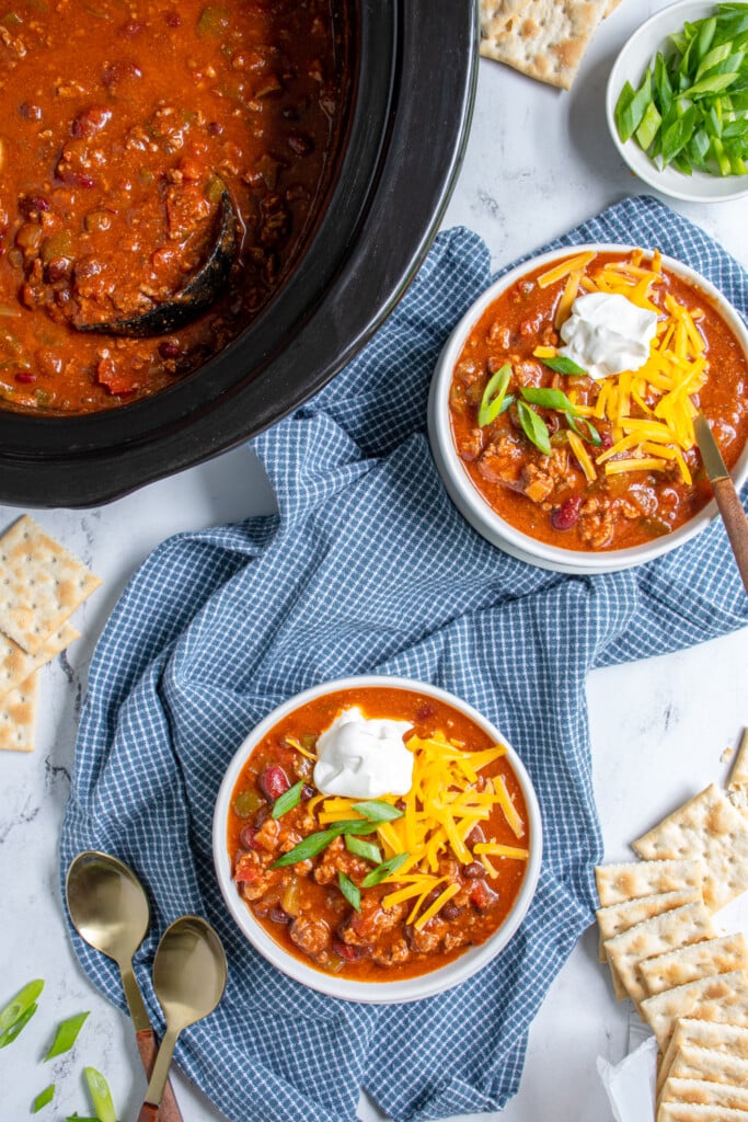 Overhead view of a tabletop with a crockpot full of chili and two bowls of chili next to it with garnishes, spoons, and saltine crackers.