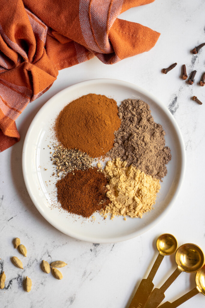 Overhead view of a small white plate with five little piles of spices that make up chai spice mix.