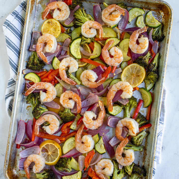 An angled sheet pan on a kitchen towel holding baked shrimp and veggies.