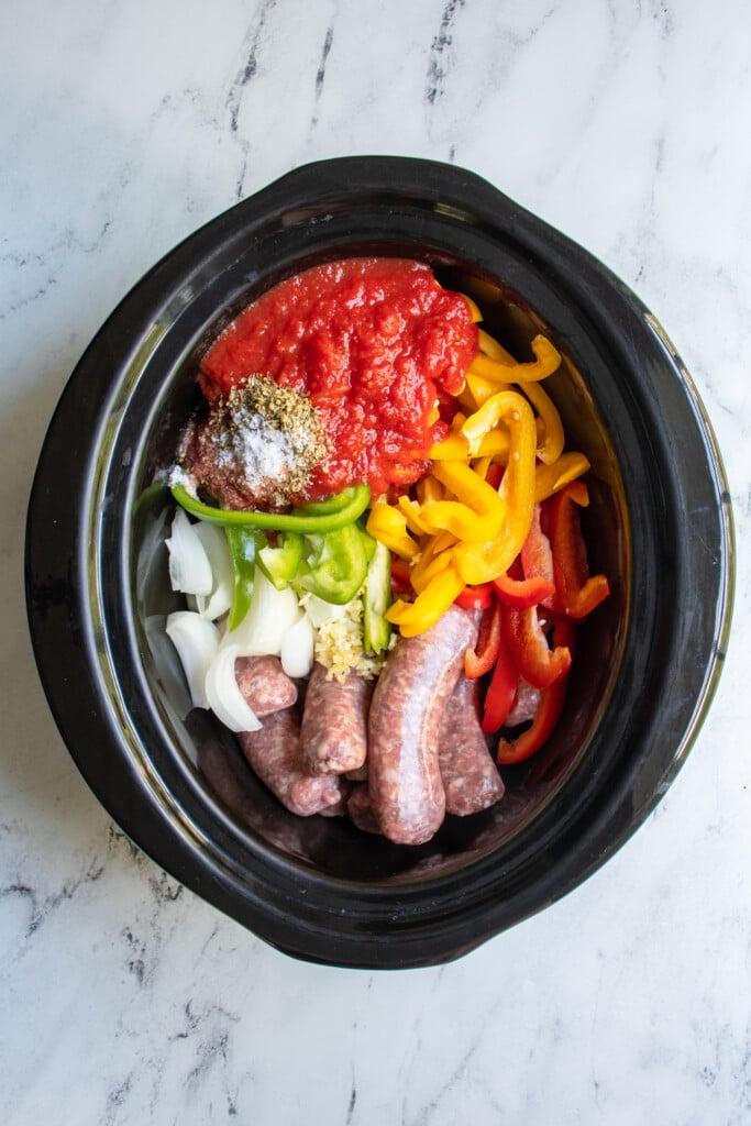 Overhead view of a crockpot with uncooked ingredients for sausage and peppers including Italian sausages, sliced bell pepper, and crushed canned tomatoes.