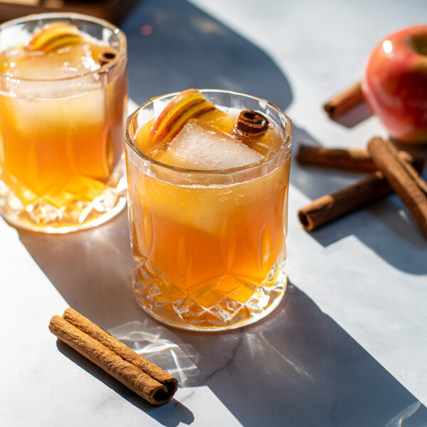 Hard sun light hitting two nice whiskey glasses full of apple cider bourbon cocktail garnished with cinnamon sticks and apple slices.