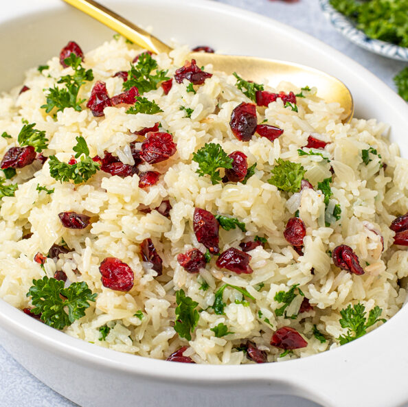 A white serving dish with a gold spoon in white rice pilaf with dried cranberries and fresh parsley.