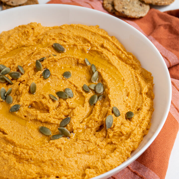 A large white bowl filled with orange colored pumpkin hummus topped with olive oil and pumpkin seeds.