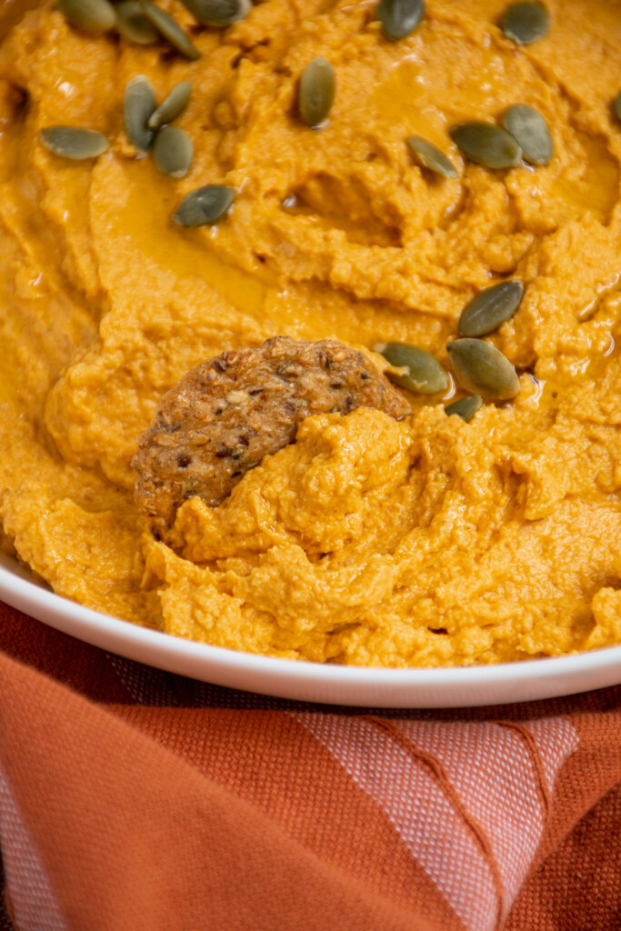 Close up of orange pumpkin hummus in a bowl being scooped out by a cracker.