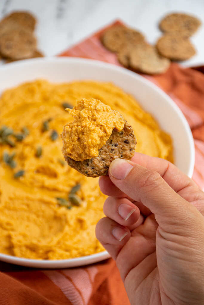 A hand holding up a round cracker hand covered with orange savory pumpkin hummus.