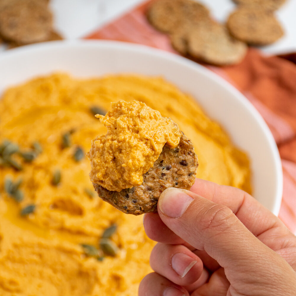 A hand holding up a round cracker hand covered with orange savory pumpkin hummus.