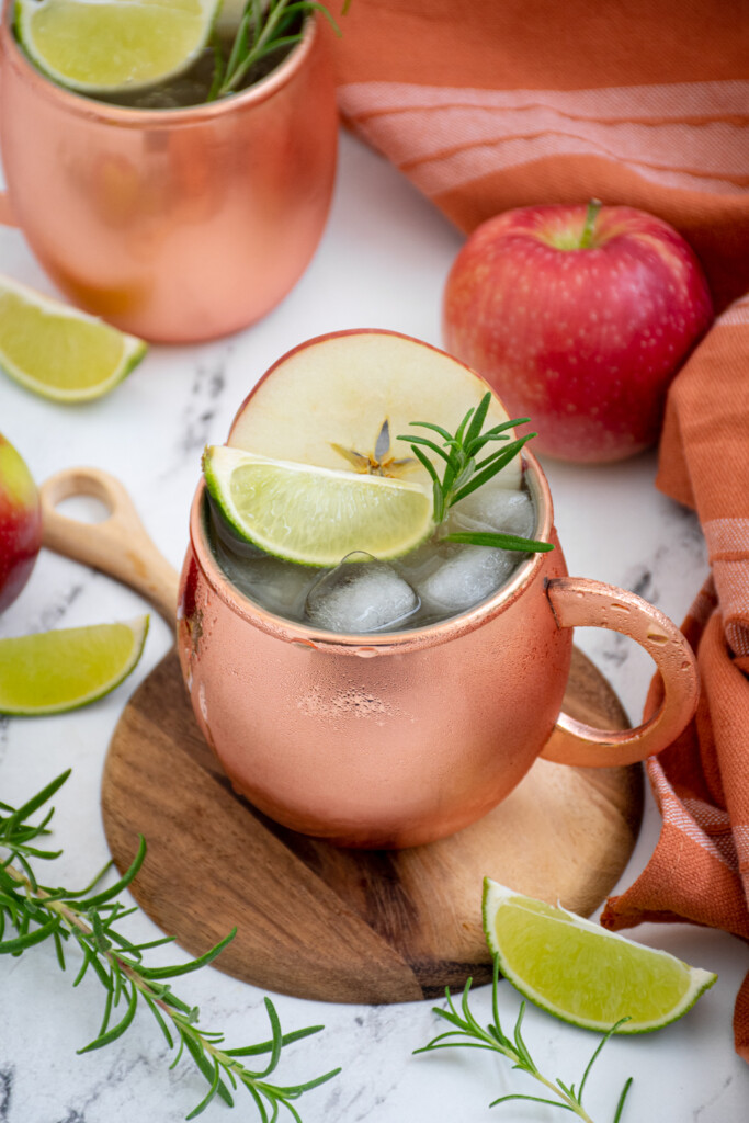 A copper mug on a cutting board filled with ice and an apple cider Moscow mule with an apple slice, lime wedge, and rosemary spring as garnish.