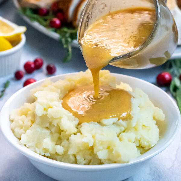 Turkey Gravy From Pan Drippings in a small glass creamer being poured onto a huge bowl of mashed potatoes, the gravy forming a large pool in the center.