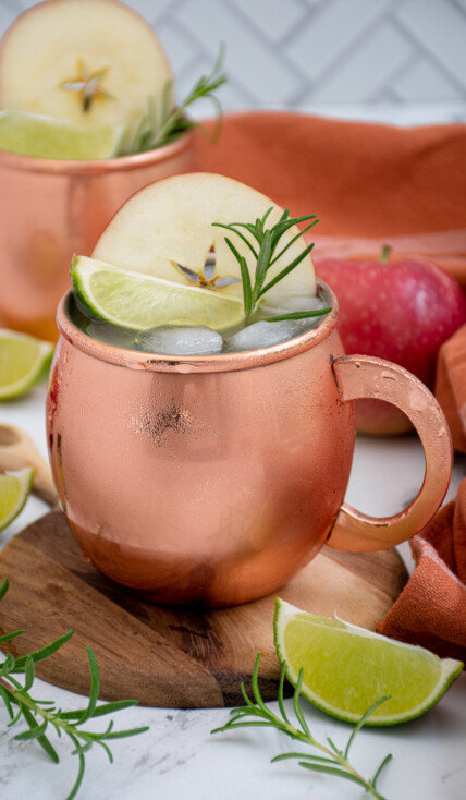 A copper mug filled with apple cider Moscow mule garnished with an apple wheel, lime wedge, and rosemary sprig.