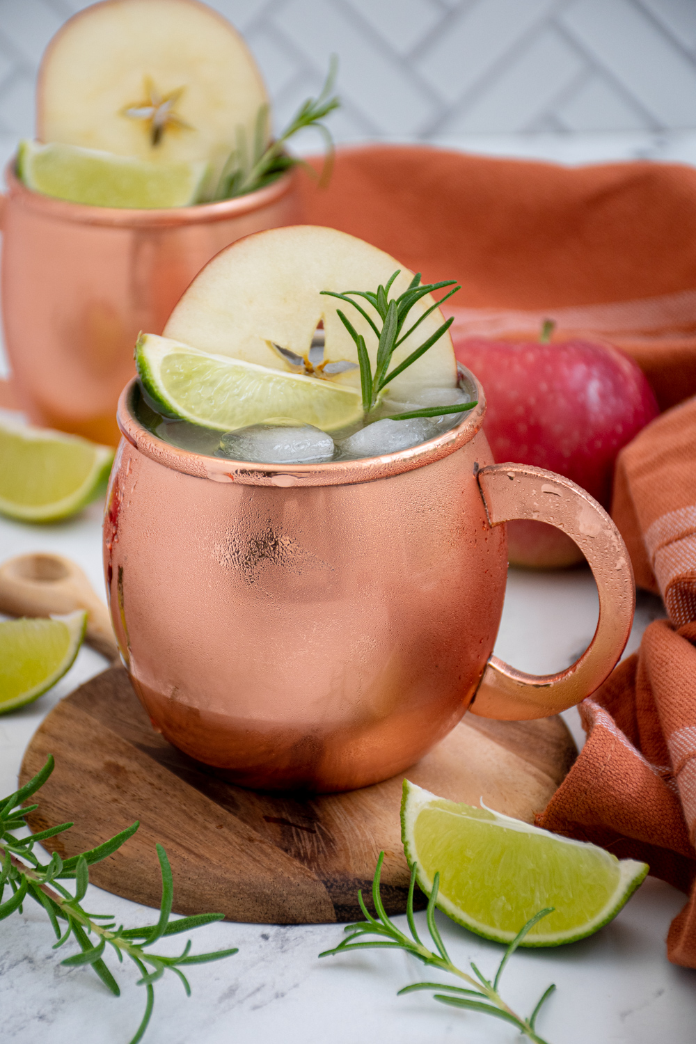 A copper mug filled with apple cider Moscow mule garnished with an apple wheel, lime wedge, and rosemary sprig.