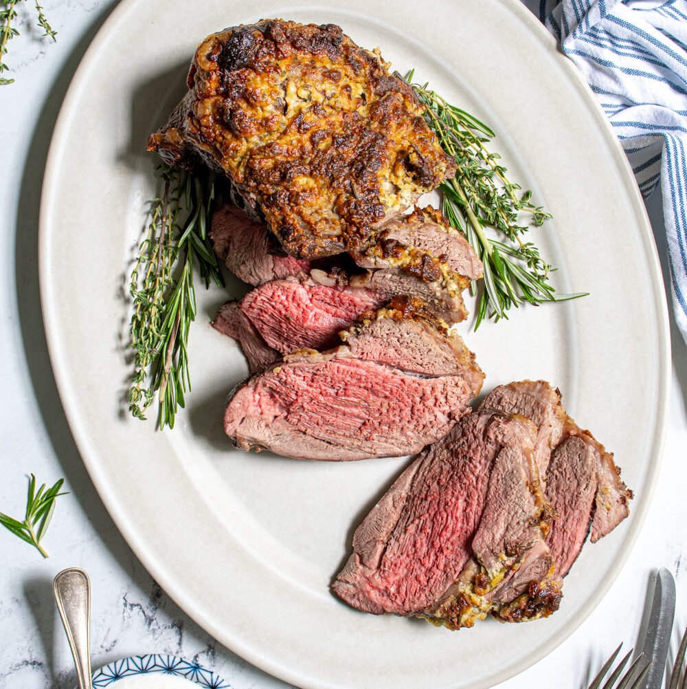 Overhead view of a roasted boneless leg of lamb on a platter, half of it sliced open revealing a tender pink inside.