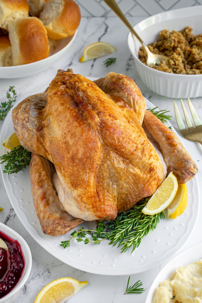 Easy Thanksgiving Turkey roasted and on a platter surrounded by herb garnishes and lemon wedges, showing off a golden brown skin.