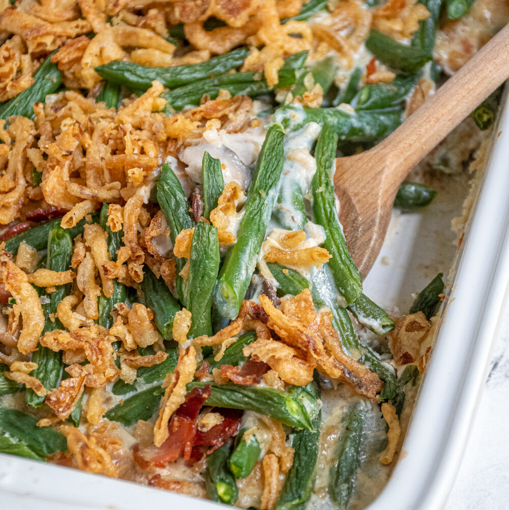 A white baking dish with green bean casserole with bacon with a serving spoon scooping some out.