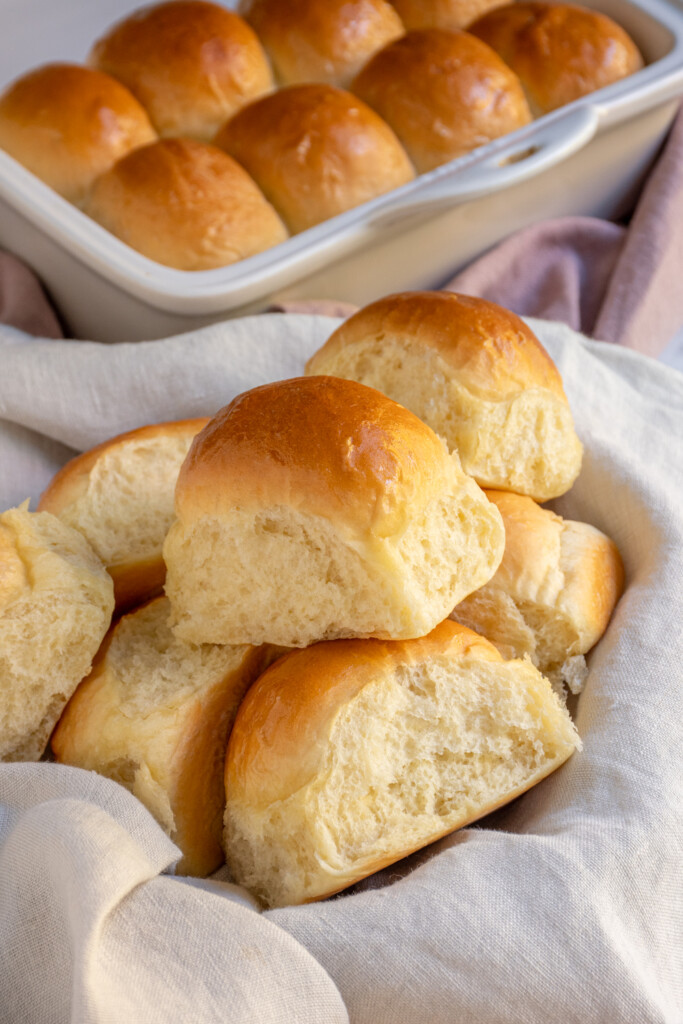 A linen lined basket filled with a bunch of golden brown soft dinner rolls.